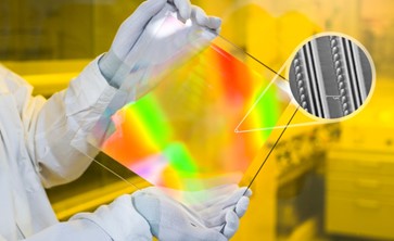 Eine Person mit weißen Handschuhen hält eine viereckige Glasplatte in die Kamera. Die Glasplatte ist in großen Teilen in grünen, roten und gelben Schimmer zu sehen. Anhand einer Einblendung wird die Nanostruktur der bunten Oberfläche im Querschnitt gezeigt.