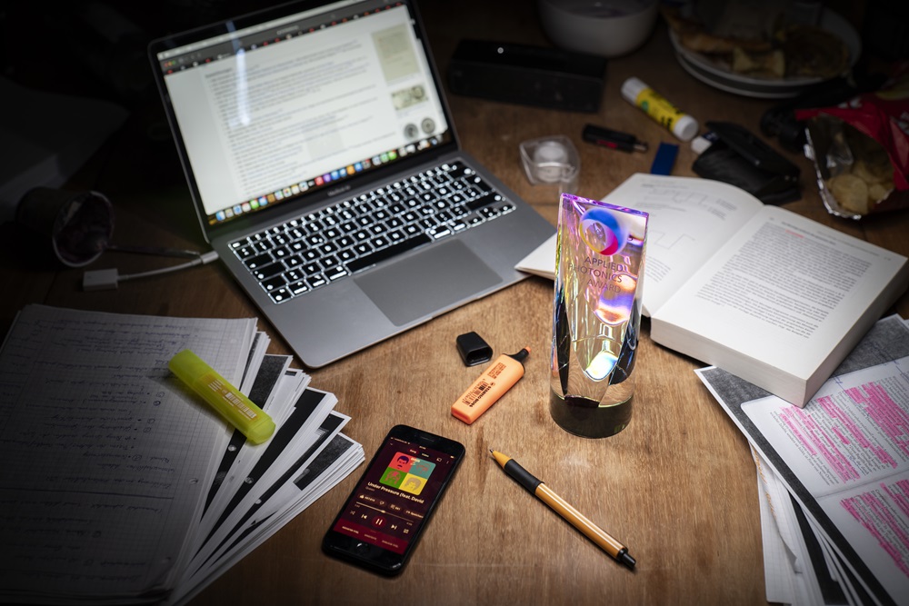 Ein Glaspokal steht auf einem Schreibtisch, umringt von Arbeitsmaterialien wie Büchern, Stiften, einem Computer und einem Smartphone.