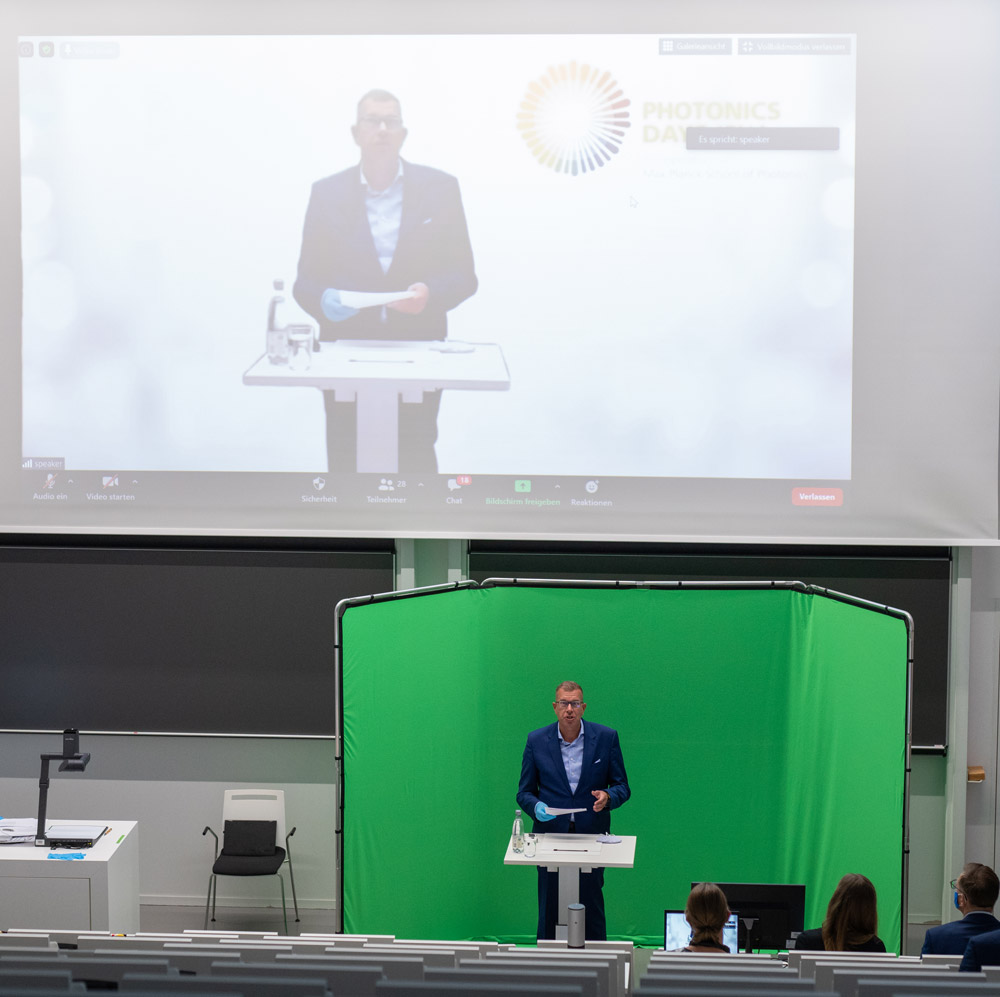 Prof. Andreas Tünnermann spricht in einem Hörsaal vor einem Green-Screen stehend zu dem Online-Publikum.