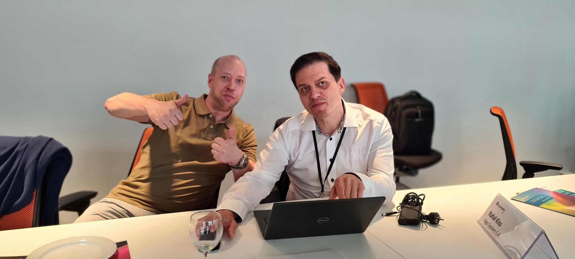 Zwei Männer sitzen gemeinsam am Computer und geben Daumen hoch in die Kamera beim Photonics Manager Compact 2022.