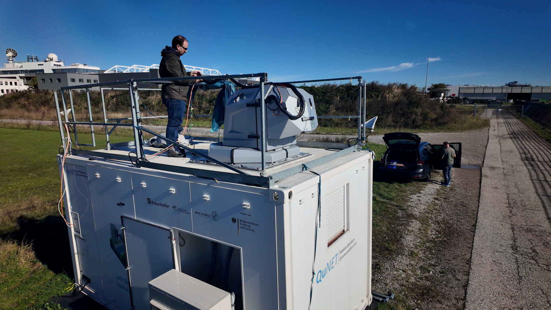 Ein weißer Container steht auf einer freien Wiese. Auf dem Dach des Containers steht ein Mann, der an der ebenfalls auf dem Dach montierten Empfangseinheit arbeitet.