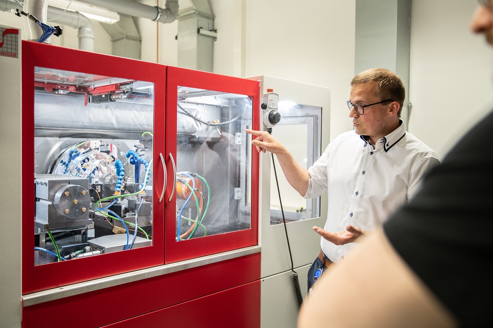 Dr. Jan Kinast stands in front of a machine and explains the process of ultra-precise free-form machining to the guests.