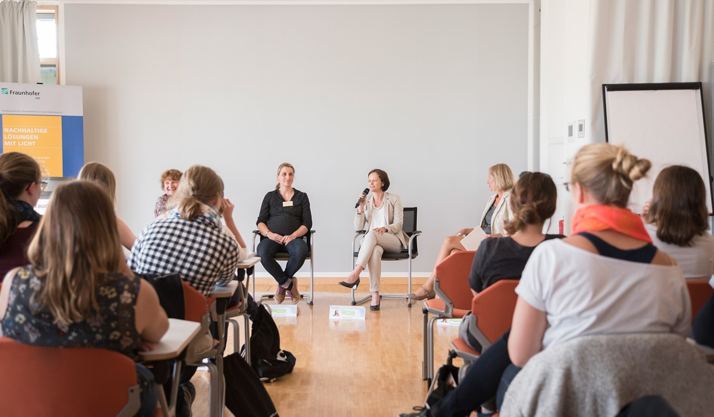 Women seated from behind. A panel discussion on career paths at Fraunhofer IOF is taking place in a brightly lit hall.