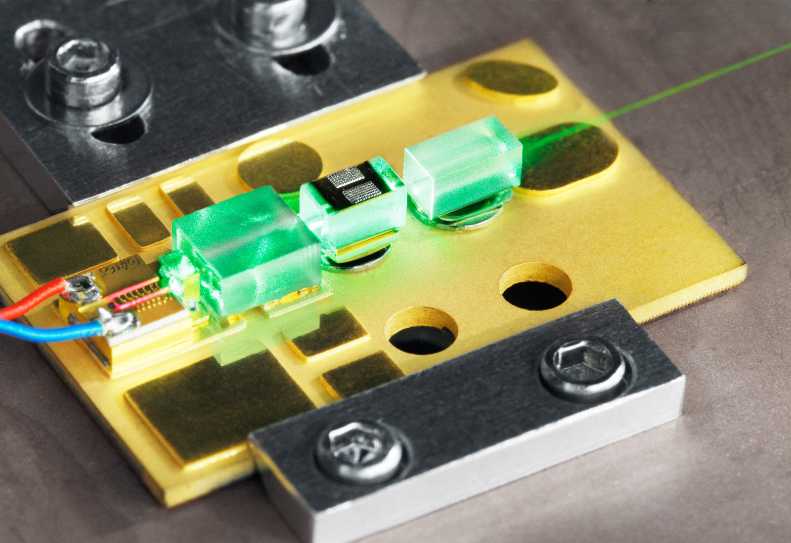 Microchip laser for use in spectroscopy: a golden plate with holes. On it are three bright green, glowing rectangles. Current is passed through the left rectangle. This is converted into a laser beam, which is passed through all three rectangles.