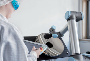 Analysis of nano- and microstructures, roughness analysis, scanning electron microscopy, X-ray microanalysis: a person is holding a tablet. In front of her is a device with a kind of gripper arm. At the end of the arm is a sensor that analyzes the surface of a concave metal mirror.