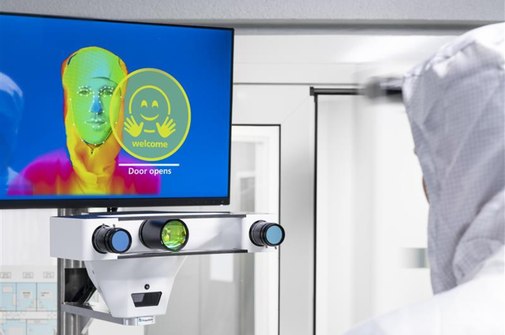 3D-Sensing, Spectral / multimodal Sensing, Image processing &amp; AI: a person stands in front of a technological device with 3 lenses. A screen can be seen above the device on which the face and the upper body of the person is shown in different colors.