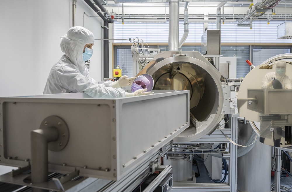 A woman in a white protective gown and mask stands in a laboratory and bends over a large machine. In her hand she holds a round mirror in which her face is reflected.