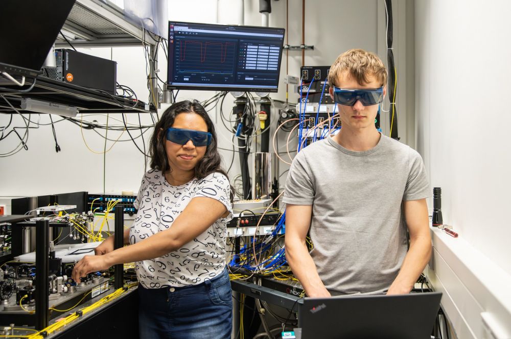 Two people are standing in a room. They are both wearing safety goggles and working.