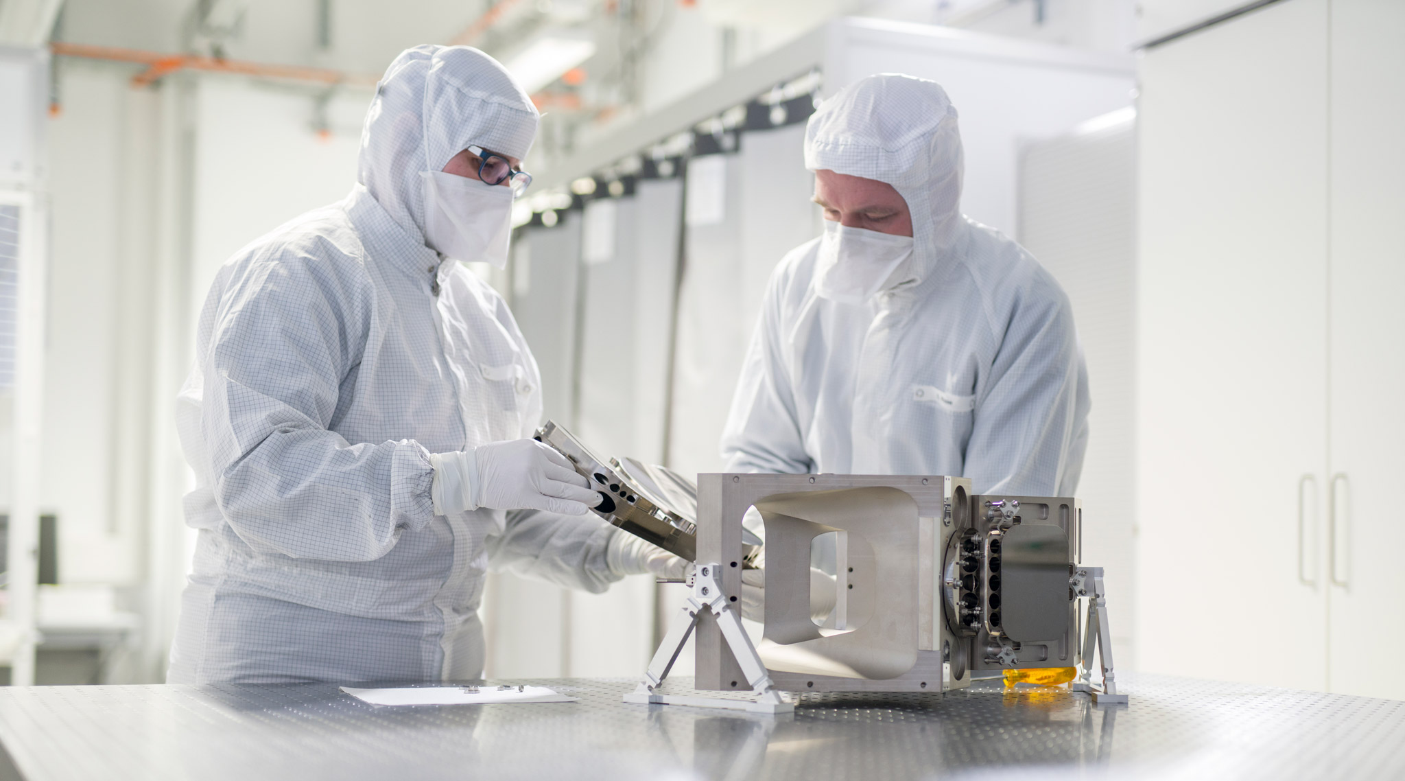 Two researchers in cleanroom clothing assemble components of a space telescope